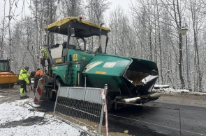 Maszyna drogowa układa asfalt na remontowanej jezdni. Obok pracują osoby w odzieży odblaskowej, a teren robót jest zabezpieczony barierkami. Wokół leży śnieg, a w tle znajdują się drzewa.
