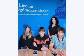 Trzech uczniów i nauczycielka pozują przy stole na tle niebieskiej ścianki z napisem o I LO w Cieszynie. Na stole leżą kalkulatory i trójwymiarowe modele brył geometrycznych z przezroczystego tworzywa.