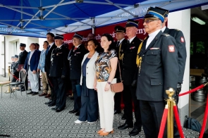 Grupa osób stoi pod namiotem podczas uroczystości. Część uczestników ma na sobie mundury, a w tle widoczny jest czerwony baner oraz niebieskie zadaszenie.