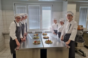 Sześciu młodych kucharzy w białych uniformach i czapkach stoi po obu stronach stalowego blatu, na którym ustawiono talerze z kolorowo podanymi daniami. Tło stanowi jasna kuchnia z oknami i profesjonalnym sprzętem.