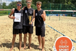Trzech chłopców w sportowych strojach stoi boso na piaskowym boisku do siatkówki plażowej. Jeden trzyma dyplom, drugi puchar. W tle siatka, banery sponsorów i drzewa. W rogu grafika z napisem „Osuch Sport”.
