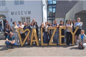 Grupa młodzieży i opiekunów pozuje przed budynkiem muzeum. Uczestnicy stoją wokół dużej przestrzennej instalacji tworzącej napis „ALZEY”, ustawionej na brukowanym placu. W tle widoczna jest jasna elewacja oraz nowoczesna zabudowa z dużymi przeszkleniami.