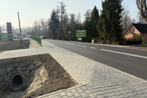 Nowa jezdnia z chodnikiem i barierką przy drodze, obok rów z przepustem, w tle stacja paliw i zabudowania.
