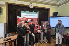 Grupa osób stojących na scenie przed banerem szkoły, część z nich trzyma nagrody i torby, a obok mężczyzna z mikrofonem prowadzi wydarzenie.