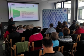 W przestronnej sali konferencyjnej grupa osób siedzi na drewnianych krzesłach, zwrócona w stronę ekranu projekcyjnego. Kobieta w zielonej bluzce prowadzi prezentację dotyczącą usług rynku pracy. Na ekranie widoczne są informacje o poradnictwie zawodowym. W tle duże okna wpuszczają naturalne światło, a na ścianie znajduje się baner z logo ratownictwa medycznego.