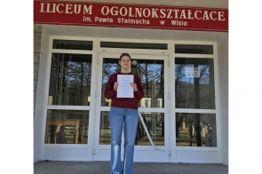 Dziewczyna stoi przed wejściem do liceum i trzyma w rękach dyplom, nad drzwiami widnieje napis z nazwą szkoły w Wiśle.