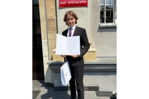 Młody chłopak stoi przed budynkiem  i prezentuje dyplom oraz trzyma białą torbę. Na elewacji budynku widoczna jest tablica z napisem „Sąd Apelacyjny".