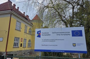 Przed żółtym budynkiem szkoły stoi duża tablica z informacją o dofinansowaniu z Funduszy Europejskich, obok widać drzewo i ogrodzenie.