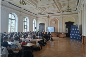 Uczniowie siedzą przy ławkach w dużej sali. Z przodu znajdują się osoba prowadząca, ekran z prezentacją oraz baner „Szkoła dla ludzi z pasją”.