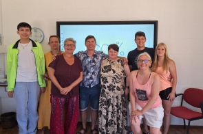 Grupowe zdjęcie dziewięciu osób stojących  w jasnym pomieszczeniu szkoleniowym. W tle widać duży ekran, a obok ścienny zegar. Osoby stoją blisko siebie, uśmiechają się do aparatu, część ma na sobie letnie, kolorowe ubrania. Wokół znajdują się czerwone krzesła i półki z materiałami.