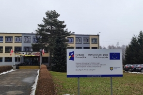 Budynek szkoły widoczny za drzewami i chodnikiem. Na trawniku przed budynkiem stoi tablica informująca o projekcie dofinansowanym przez Unię Europejską.