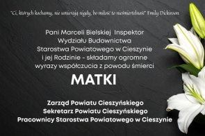 Ciemne, stonowane tło z  tekstem kondolencyjnym pośrodku, a po prawej stronie widoczny biały kwiat lilii.