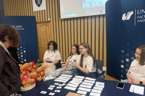 Kilka osób siedzi przy stole rejestracyjnym w sali konferencyjnej. Na stole leżą kartki z numerami, telefon, pojemniki oraz pluszowe misie. W tle widoczne są banery uczelni.