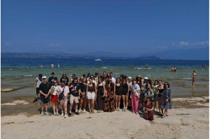 Grupa osób pozuje do zdjęcia na plaży. W tle widać wodę, kąpiących się ludzi, odległy brzeg oraz góry pod błękitnym niebem.