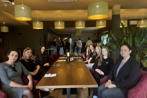 Grupa uczniów w eleganckich strojach hotelarskich ćwiczy przy recepcji. W tle widoczna jest tablica z nazwą obiektu, a atmosfera spotkania jest pełna zaangażowania i uśmiechów.