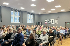 Duża grupa osób siedzi na krzesłach, zwrócona w jedną stronę, część trzyma w rękach torby lub ubrania, a niektórzy mają obok siebie laski.