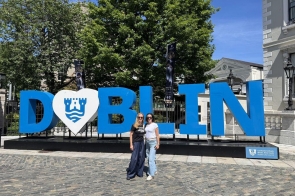 Dwie kobiety pozują do zdjęcia przy dużym napisie „Dublin” w niebiesko-białej kolorystyce. W tle widać drzewa, zabudowę miejską oraz słoneczne niebo.