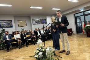 Dwie osoby stoją przy mikrofonach w sali szkolnej. W tle widać siedzącą publiczność, dekoracje kwiatowe oraz flagi.