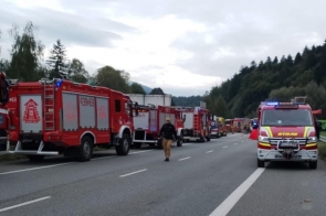 Szereg czerwonych wozów strażackich i karetek stoi na zablokowanej drodze ekspresowej, po pasie awaryjnym stoi bus straży z włączonymi światłami. Między pojazdami idą ratownicy, a w tle widać drzewa i pagórki pod pochmurnym niebem.