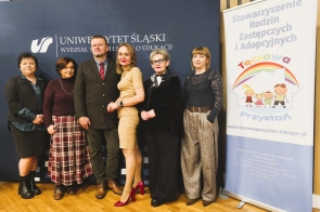 Grupa osób pozuje do zdjęcia na tle baneru Uniwersytetu Śląskiego oraz roll-upu Stowarzyszenia Rodzin Zastępczych i Adopcyjnych „Tęczowa Przystań”.