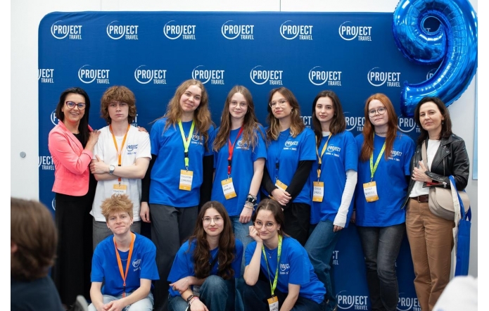 Grupa młodzieży w niebieskich koszulkach z logo „Project Travel” pozuje na tle ścianki z nazwą projektu. Wśród nich dwie kobiety w eleganckich stylizacjach, kolorowe identyfikatory na szyjach i duży balon w kształcie cyfry 9.