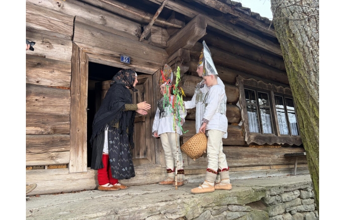 Kobieta stoi w drzwiach drewnianego domu i rozmawia z trójką dzieci ubranych w stroje ludowe. Jedno z nich trzyma ozdobioną gałązkę, a inne koszyk. W tle widoczna jest ściana chaty oraz okno.