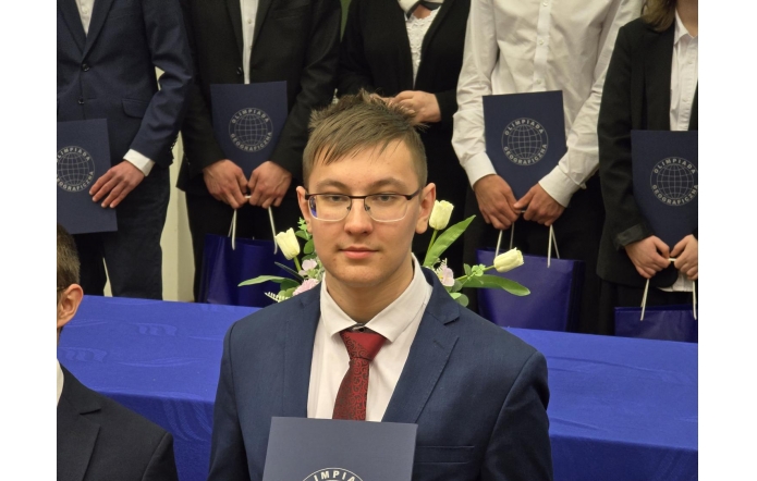 Uczeń w okularach i garniturze z czerwonym krawatem trzyma teczkę z logo Olimpiady Geograficznej. W tle grupa młodzieży w odświętnych strojach oraz stół udekorowany białymi tulipanami. Uroczysta atmosfera akademickiego wydarzenia.