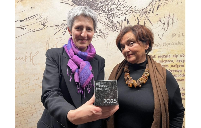 Dwie kobiety pozują do zdjęcia. Po lewej stronie stoi Irena French Dyrektor Muzeum Śląska Cieszyńskiego, która pokazuje wyróżnienie do zdjęcia,a po prawej Janina Żagan Starosta Cieszyński. W tle widoczna jest ściana z grafikami i napisami.