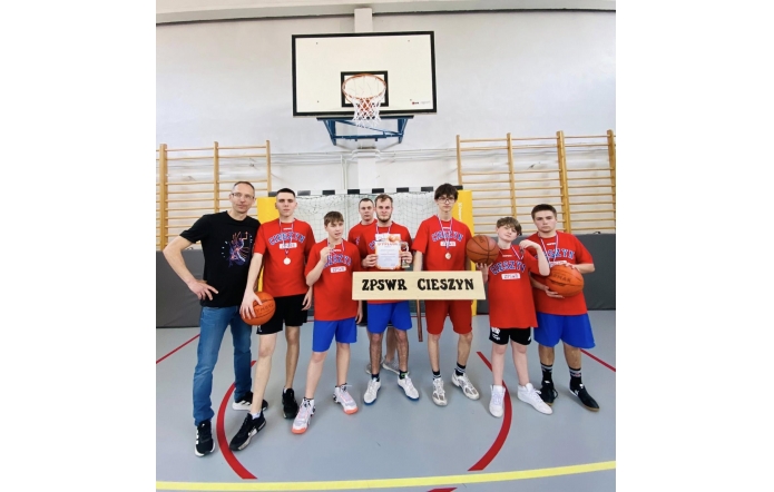 Grupa chłopców w czerwonych koszulkach „Cieszyn” stoi w sali gimnastycznej z medalami, piłkami i dyplomem. Obok nich stoi trener, a z przodu widoczna jest tabliczka „ZPSWR Cieszyn”.