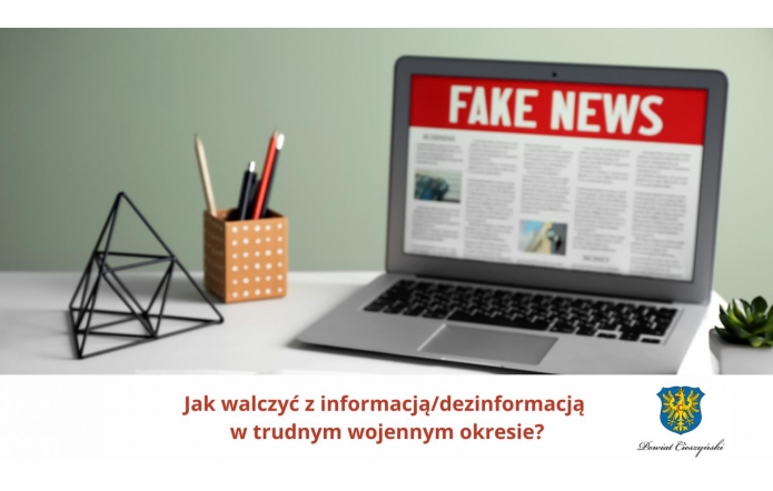 Na ekranie laptopa widoczny nagłówek „FAKE NEWS” na czerwonym tle, sugerujący tematykę fałszywych informacji. Obok komputerowego ekranu stoi pojemnik z długopisami i ołówkami oraz geometryczna dekoracja. U dołu grafiki widnieje pytanie: „Jak walczyć z informacją/dezinformacją w trudnym wojennym okresie?” oraz herb Powiatu Cieszyńskiego.