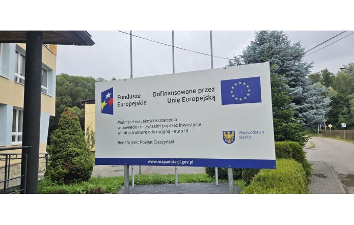 Tablica informacyjna o projekcie dofinansowanym przez Unię Europejską. Widoczne są logotypy Funduszy Europejskich, Unii Europejskiej i Województwa Śląskiego, nazwa projektu dotyczącego inwestycji w infrastrukturę edukacyjną oraz informacja, że beneficjentem jest Powiat Cieszyński.