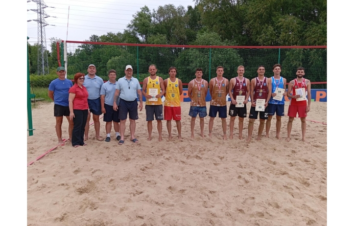 Grupa osób stoi na boisku do siatkówki plażowej. Pośrodku uśmiechnięci zawodnicy w sportowych strojach prezentują medale i dyplomy. Obok nich organizatorzy w koszulkach polo. W tle siatka i drzewa, atmosfera sportowego święta.