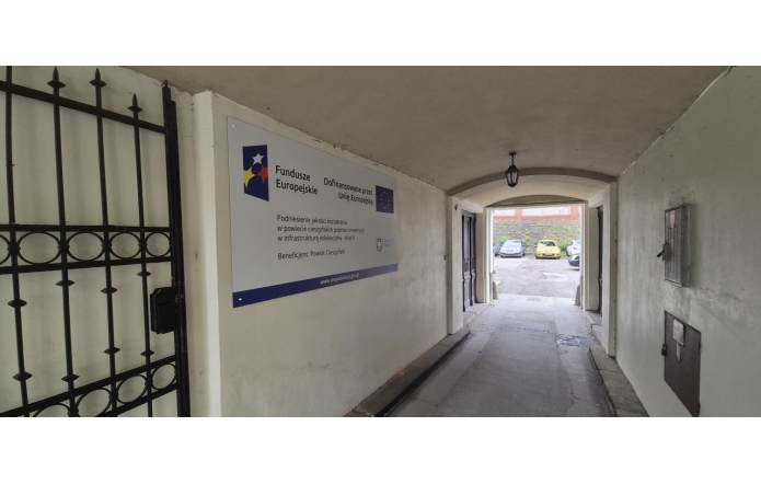 Korytarz przejściowy w budynku z tablicą informacyjną o projekcie dofinansowanym przez Unię Europejską. Po lewej stronie widoczna jest metalowa krata, a w głębi przejście prowadzące na zewnątrz.