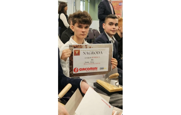 Dwóch młodych mężczyzn siedzi na sali i jeden z nich trzyma dyplom z napisem „Nagroda za zajęcie III miejsca” w turnieju budowlanym. Na dokumencie widoczna jest informacja o bonie o wartości 400 zł oraz logo firmy.