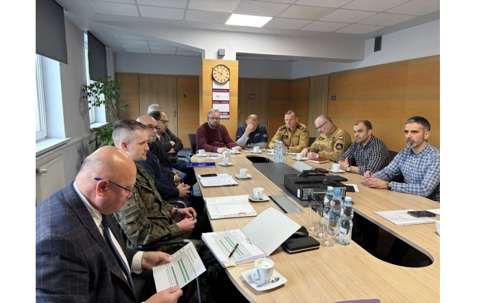 W sali konferencyjnej kilkunastu mężczyzn siedzi przy dużym stole podczas spotkania. Na stole leżą dokumenty, laptopy, filiżanki z kawą i butelki wody. Część uczestników ma na sobie mundury, a pozostali ubrani są w stroje cywilne.