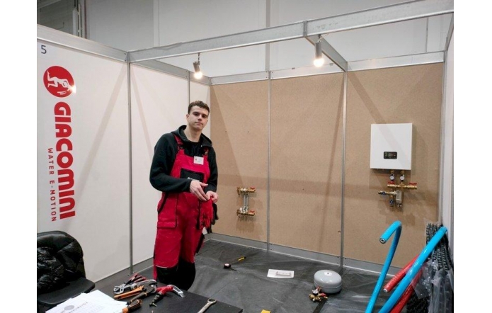 W przestrzeni targowej lub wystawowej wydzielono stanowisko techniczne z logotypem marki związanej z systemami wodnymi. Metalowa konstrukcja z tablicami w tle wyznacza obszar pracy. Młody mężczyzna w czerwonym kombinezonie roboczym i czarnym polarze trzyma narzędzie, stojąc przy stanowisku montażowym z rurami i elementami hydraulicznymi. Wokół rozłożone są narzędzia i dokumentacja. 