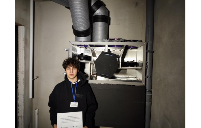 Młody mężczyzna z kręconymi, ciemnymi włosami stoi w wąskim pomieszczeniu technicznym przed otwartą centralą wentylacyjną. Ma na sobie czarną bluzę z kapturem oraz identyfikator na niebieskiej smyczy i trzyma w dłoniach kartkę z dokumentem. Za nią widoczna jest metalowa obudowa urządzenia z odsłoniętymi filtrami i wentylatorami, a nad nim biegną duże, szare rury wentylacyjne przymocowane do ściany.