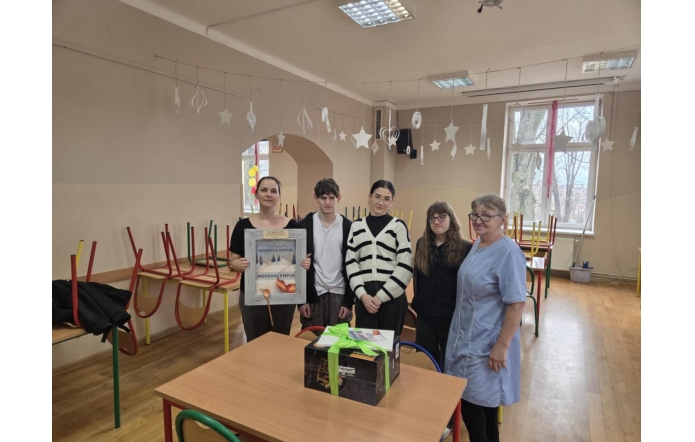 W sali lekcyjnej pięć osób stoi za stołem. Na blacie znajduje się duże pudełko przewiązane zieloną wstążką. Jedna z kobiet trzyma plakat z napisem o „Najlepszych beskidzkich kreplikach”. W tle widać podniesione krzesła ustawione na ławkach oraz okna i wiszące dekoracje w kształcie gwiazdek.