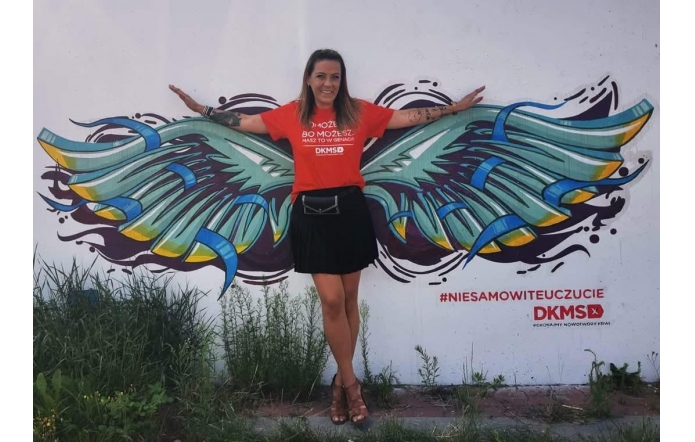   Uśmiechnięta Marta Tabaka w czerwonej koszulce z logo DKMS pozuje z rozpostartymi ramionami na tle ściennego muralu z barwnymi skrzydłami, tworząc iluzję, jakby sama je posiadała; obok widoczny hasztag #niesamowiteuczucie.