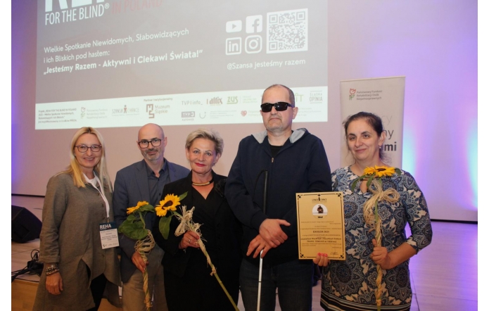 Pięć osób stoi na tle ekranu z logo konferencji REHA FOR THE BLIND IN POLAND. Troje z nich trzyma słoneczniki, jedna osoba trzyma dyplom. Wszyscy są uśmiechnięci, ubrani elegancko. W tle wyświetlone są logotypy partnerów wydarzenia