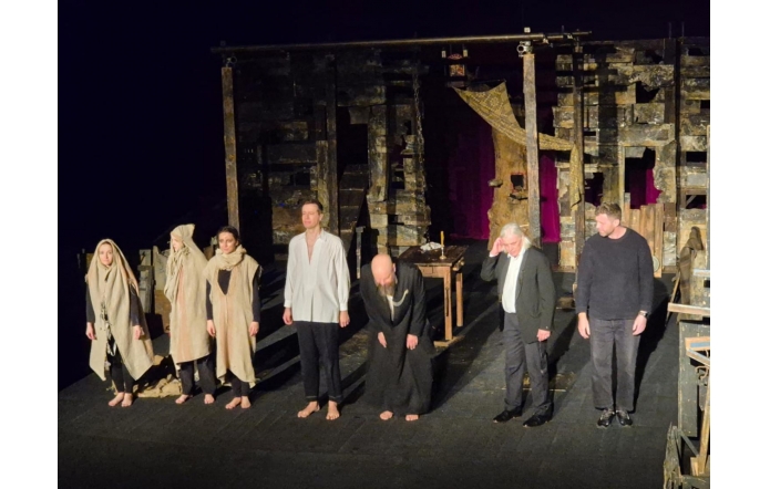 Aktorzy stoją na scenie po zakończeniu spektaklu. W tle widoczna jest teatralna scenografia, a część osób ubrana jest w proste, jasne kostiumy.