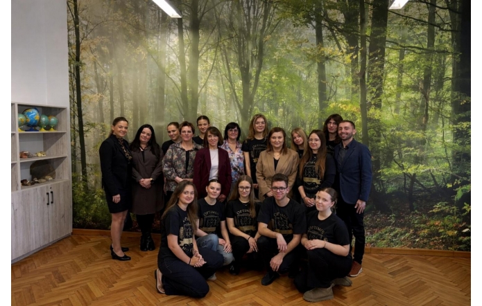 Grupa osób stoi i klęczy do wspólnego zdjęcia w pomieszczeniu z leśną grafiką na ścianie, obok widać regał z globusami.