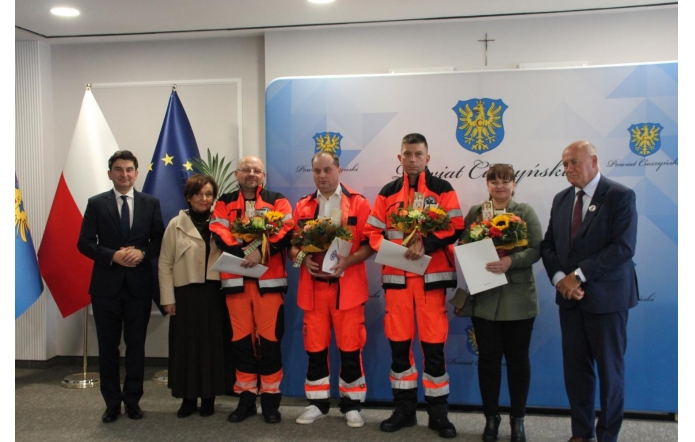 Siedem osób stoi przed ścianką z herbem Powiatu Cieszyńskiego. Trzech ratowników medycznych w pomarańczowych strojach trzyma bukiety kwiatów i dokumenty. Po bokach stoją przedstawiciele władz, a w tle widoczne są flagi Polski i Unii Europejskiej.