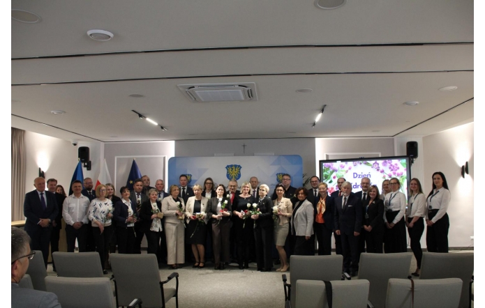 Duża grupa osób stoi ustawiona w kilku rzędach do wspólnego zdjęcia na tle ścianki z herbem. Część osób trzyma w rękach kwiaty i niewielkie pudełka.