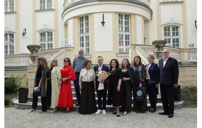 Grupa osób stoi przed pałacem na schodach wejściowych i pozuje do wspólnego zdjęcia. Jedna osoba trzyma bukiet kwiatów, a w tle widoczna jest jasna elewacja budynku.