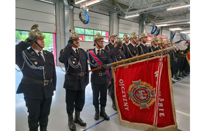 Strażacy w galowych mundurach i złotych hełmach stoją w hali podczas uroczystości.Na pierwszym planie widoczny jest czerwony sztandar strażacki oraz osoby salutujące.