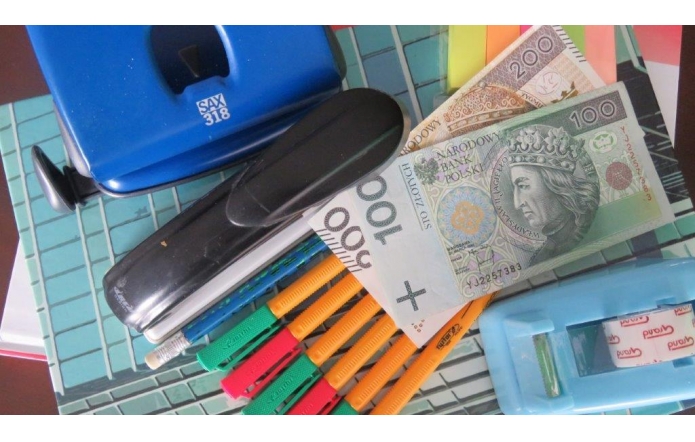 Zestaw biurowych przedmiotów ułożonych na stosie zeszytów i notesów: dziurkacz, zszywacz, kilka nożyków do papieru, długopisy, taśma klejąca w przezroczystym podajniku oraz kolorowe karteczki samoprzylepne. Pomiędzy nimi widoczne są banknoty polskie o nominałach 100 i 200 złotych.