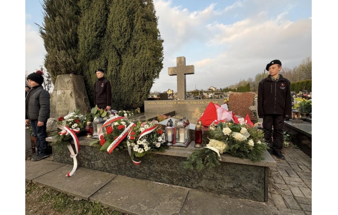 Przy grobie z krzyżem ustawiono kompozycje z biało-czerwonymi wstęgami, a sam grób ozdobiony jest wieńcami, kwiatami i zniczami. Po obu stronach stoją młodzi mężczyźni.