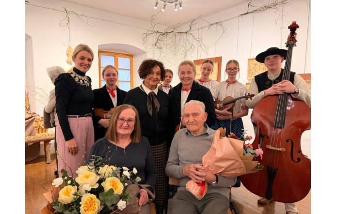 Grupa osób pozująca do wspólnego zdjęcia we wnętrzu galerii. Na pierwszym planie siedzą dwie osoby trzymające bukiety kwiatów, a za nimi stoją inni uczestnicy, w tym młodzież z instrumentami muzycznymi.