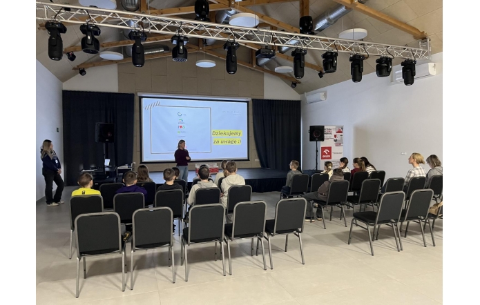 Sala konferencyjna z ustawionymi w rzędach krzesłami skierowanymi w stronę sceny. Na scenie znajduje się ekran z wyświetlaną prezentacją oraz sprzęt nagłaśniający i oświetleniowy. 